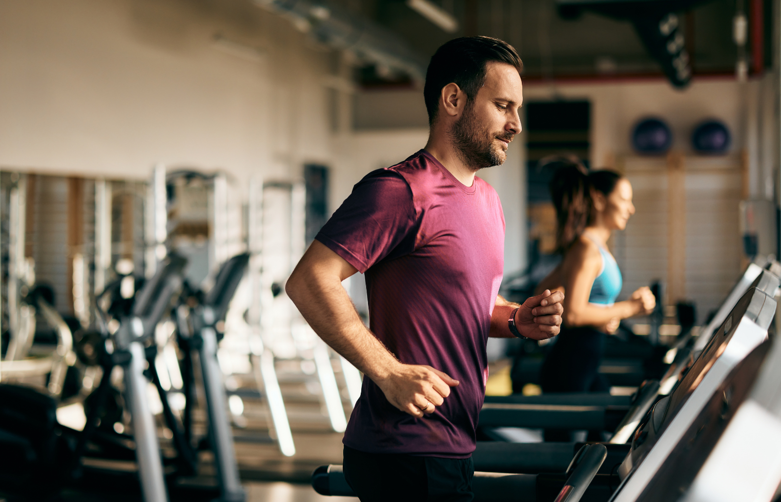 Side profile of a male and female running on a treadmill at the gym, focus on the man.