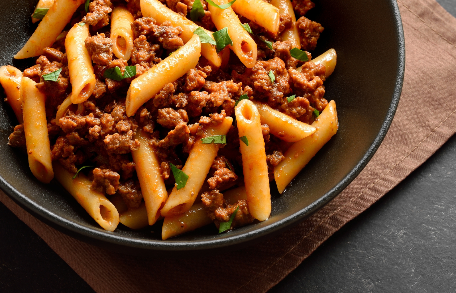 Penne pasta with mince beef in a black bowl on a brown napkin