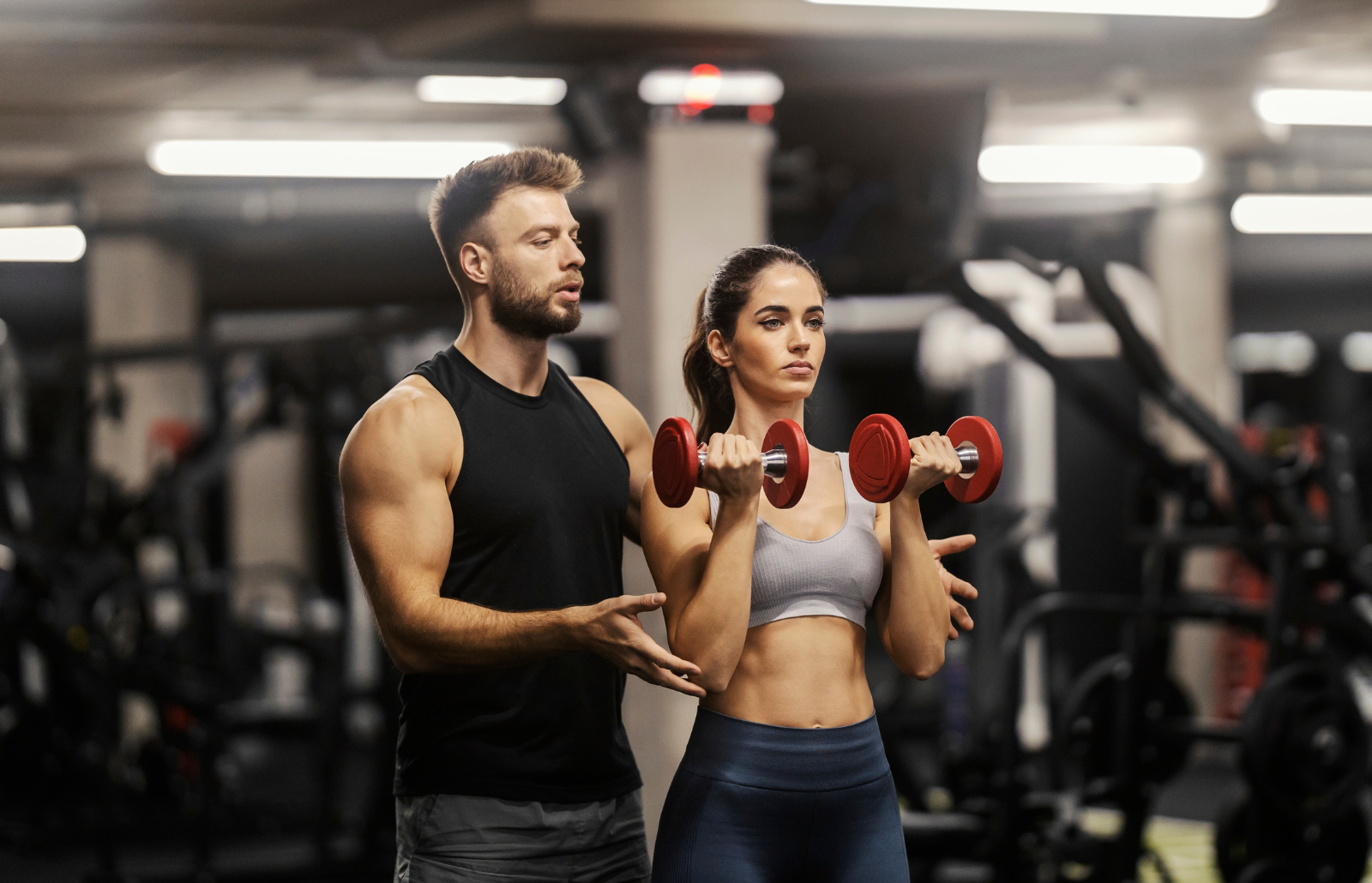 A fit sportswoman in shape is doing exercises with dumbbells in a gym and her coach is helping her.